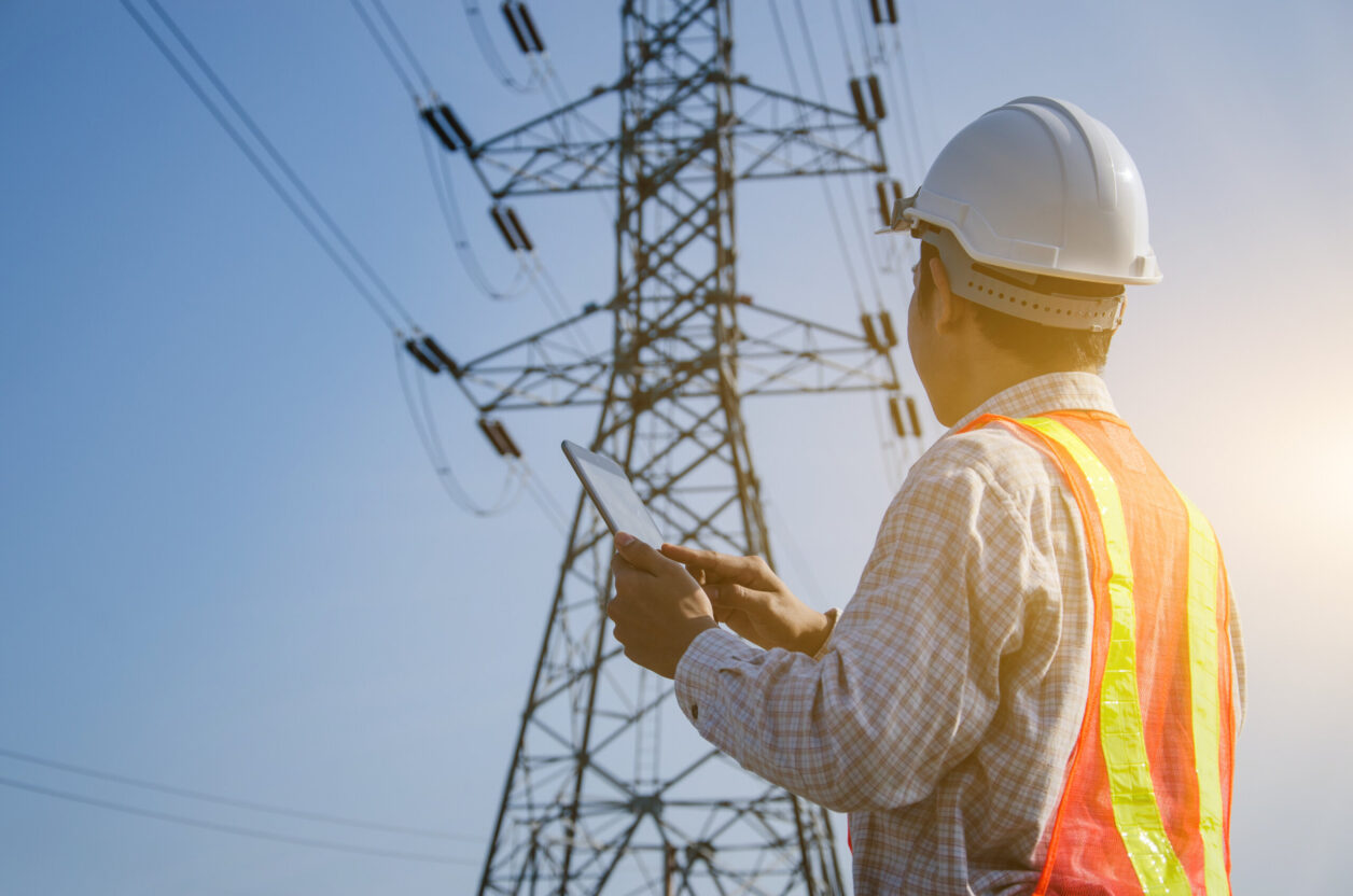 Electrical engineer surveys high voltage transmission tower from ground with tablet